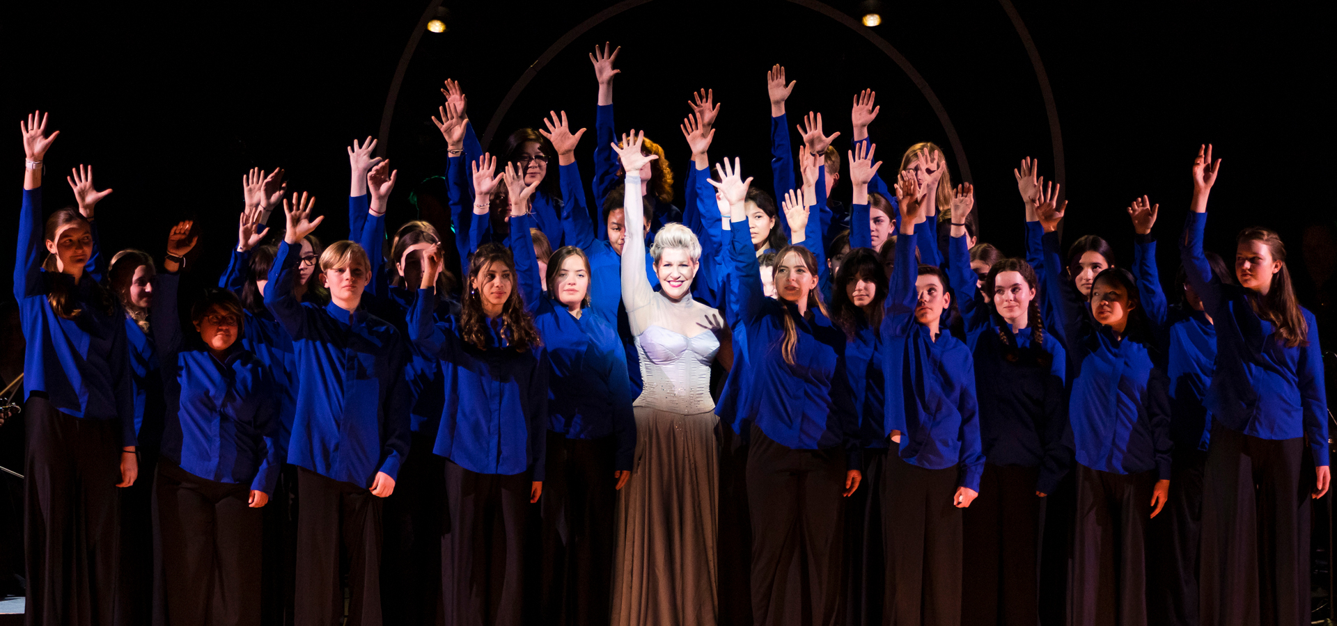 Joyce DiDonato in a white dress on stage with young people, all with hands raised and smiling