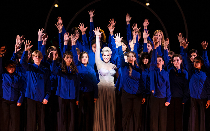 Joyce DiDonato in a white dress on stage with young people, all with hands raised and smiling