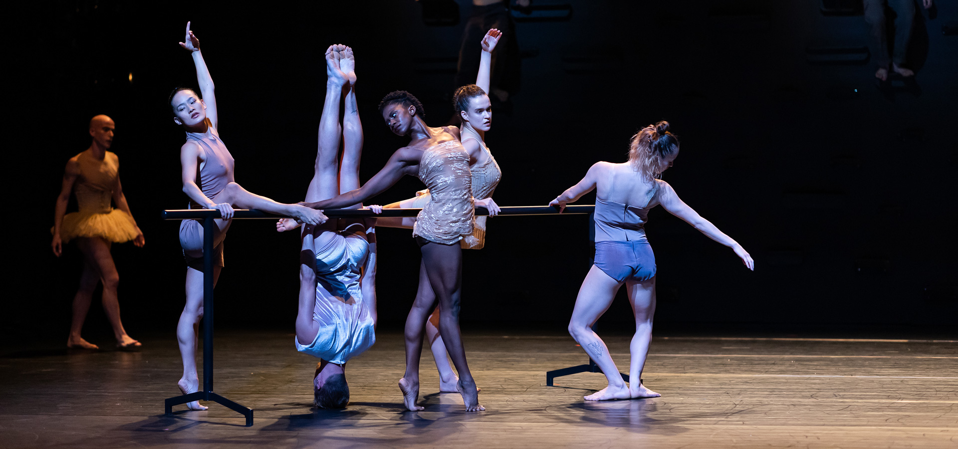 Performers in the Batsheva Dance group configure in ballet poses around a barre on the stage, one dancer balanced upside down on the crown of her head.