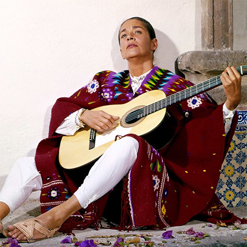 Chavela Vargas strums a guitar while wearing a traditional outfit with bright embroidered colors.