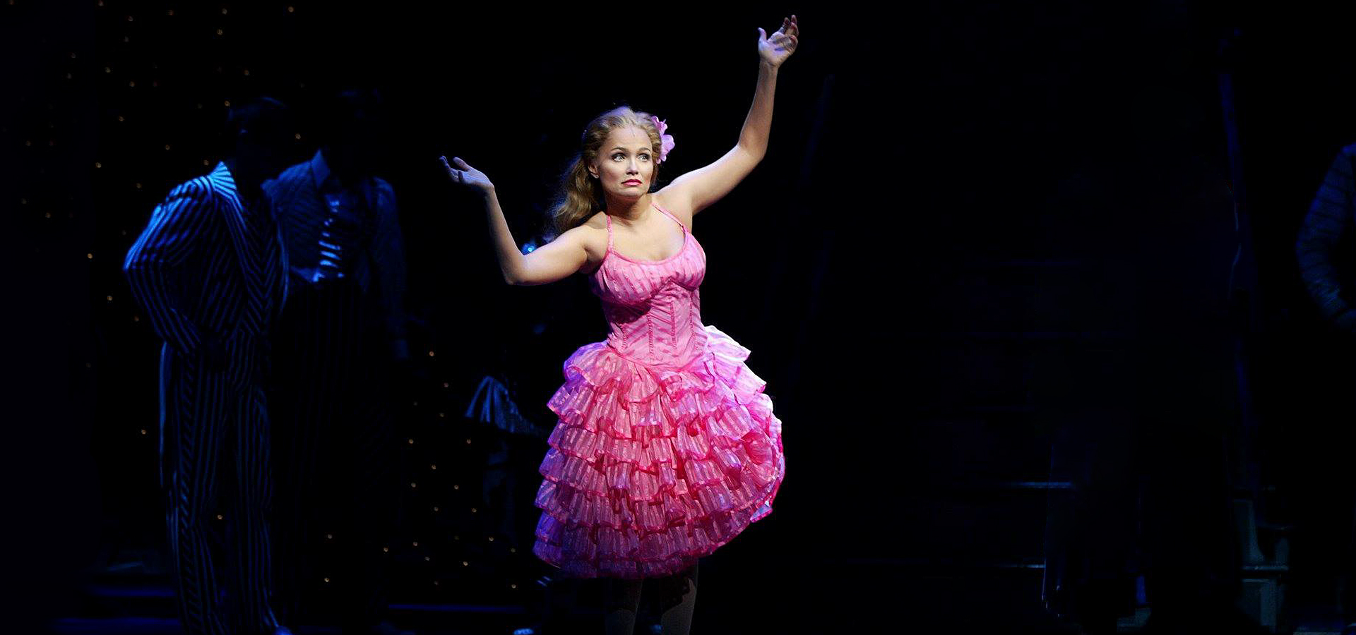 Kristin Chenowith is brightly lit on a dark stage, standing in a vibrant, ruffled hot pink dress with her arms raised.