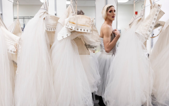 An image of white costumes hanging from a rack, with a ballerina visible in costume in the background.