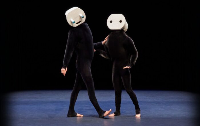 Two performers in all-black outfits with animated geometric masks on their heads stand next to each other center-stage.