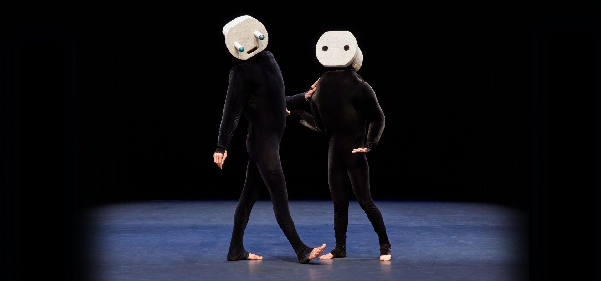 Two performers in all-black outfits with animated geometric masks on their heads stand next to each other center-stage.