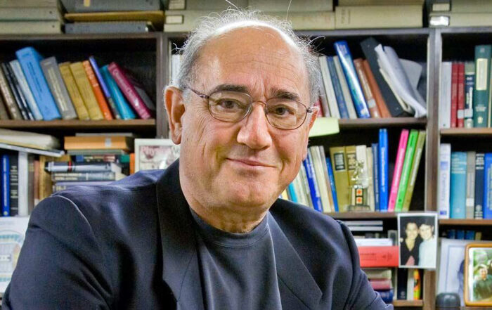 Alex Pines, an older white man with gray hair and thin round wire glasses, smiles slightly into the camera in front of full bookshelves