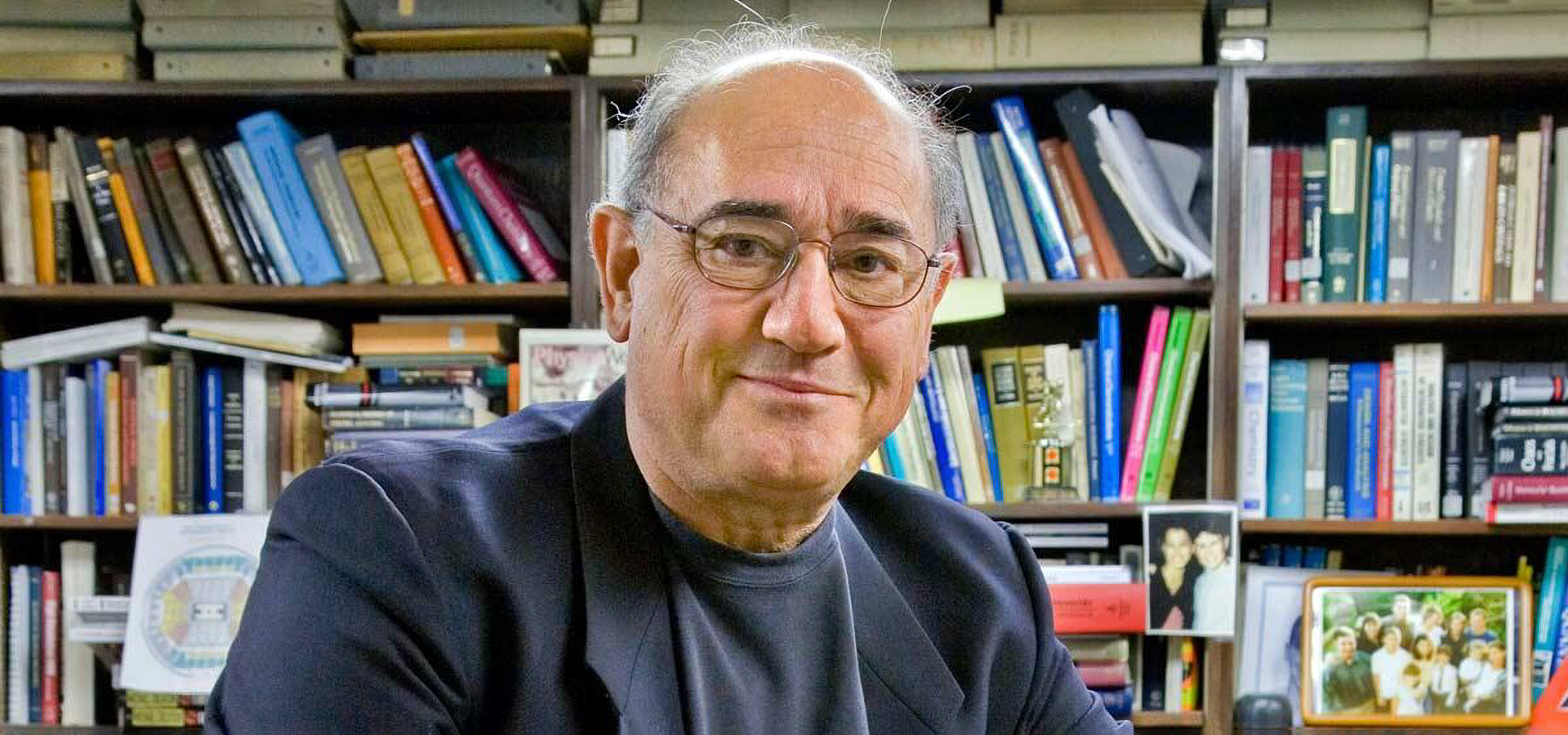 Alex Pines, an older white man with gray hair and thin round wire glasses, smiles slightly into the camera in front of full bookshelves