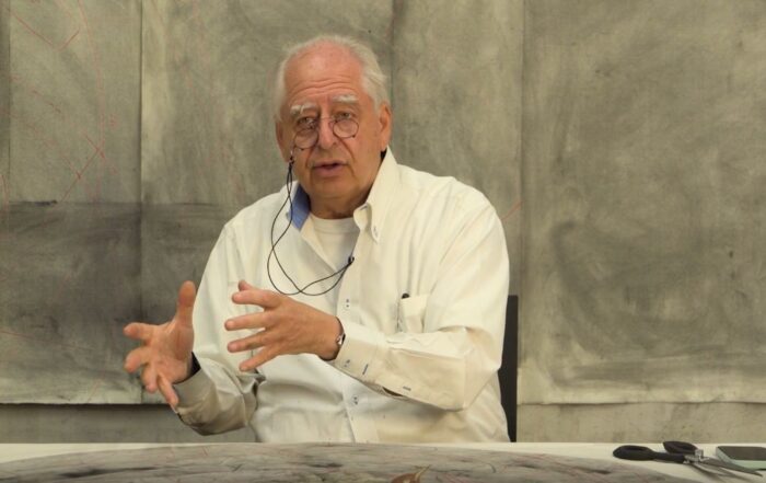 William Kentridge, an older man wearing a white dress shirt and glasses, sits at a table against a gray background and gestures with his hands.