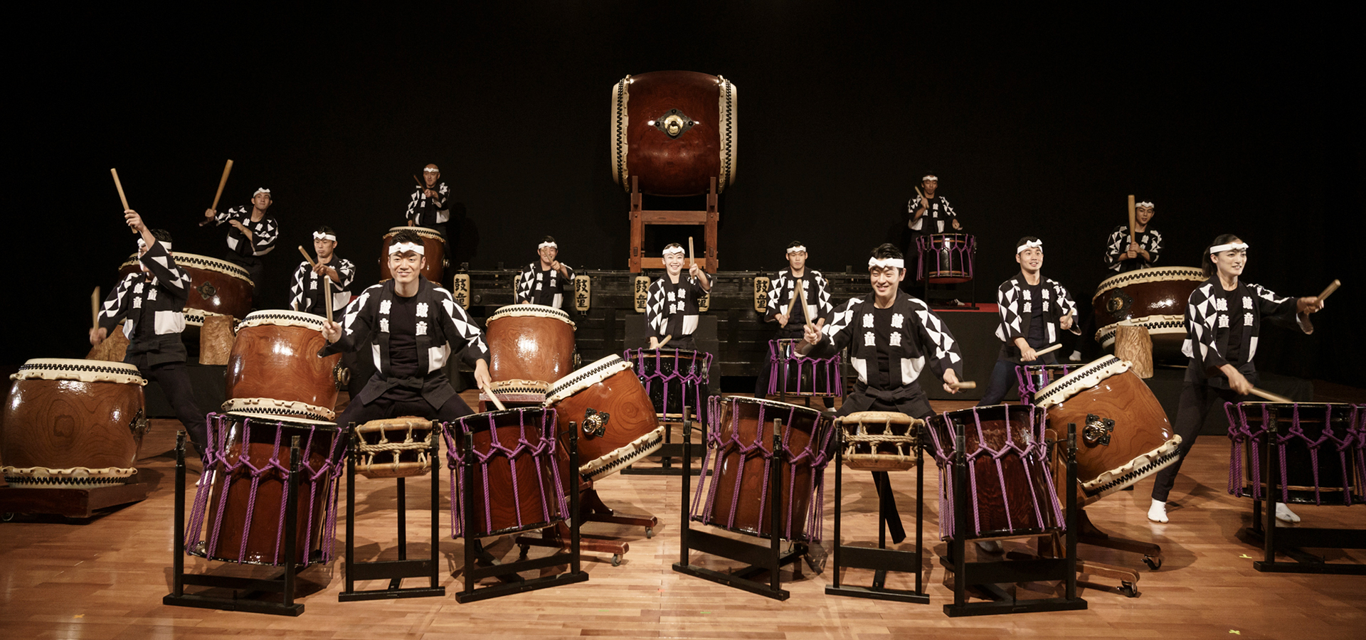 Members of the Kodo Troupe, wearing traditional Japanese attire, sit in front of numerous large drums on stage during an enthusiastic performance of Warabe.