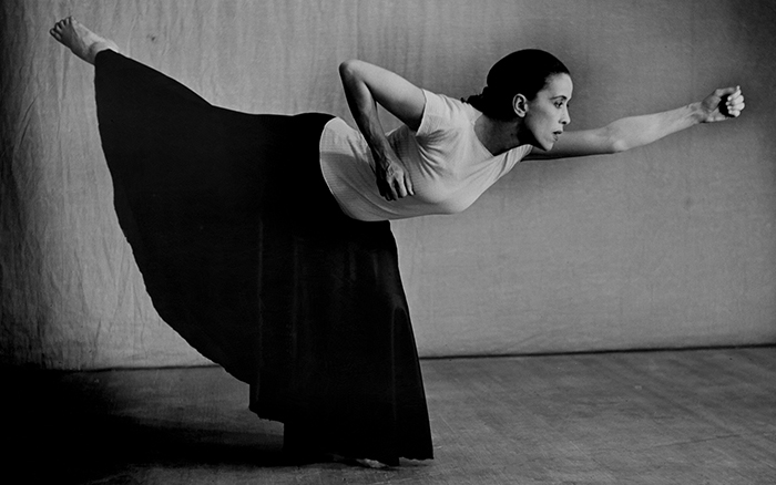 Black and white photo of dancer and choreographer Martha Graham wearing a white top and long black skirt in pose