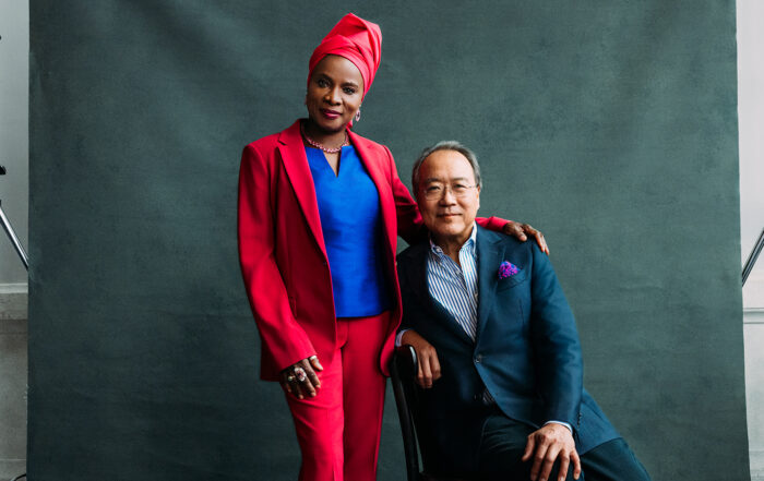 Angelique Kidjo, an African woman in a vibrant red suit and blue blouse, and Yo-Yo Ma, an Asian man in a suit, smile as they pose in front of a grey backdrop.