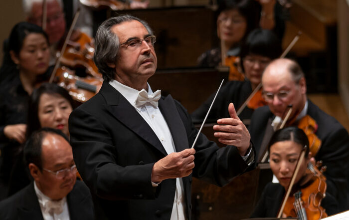Riccardo Muti stands at a podium with his baton in hand, conducting with a serious expression on his face.