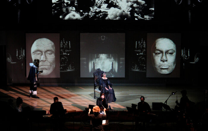 The lead actors of Opera Parallele's La Belle et La Bête perform onstage, with La Bête in the background, gazing at Belle as she sits in a chair, wearing a somber expression.