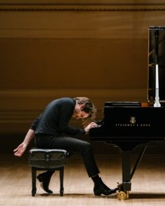 Alexandre Kantorow, a French man wearing all black, sits while playing on a grand piano with his head bowed down towards the keys.