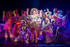 Performers gather in elaborately textured and colorful costumes adorned with fur, gemstones, and glitter; raising their hands in a group formation on stage.