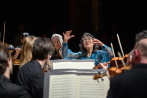 The Mahler Chamber Orchestra conducted by Mitsuko Uchida, who is shown center-stage in a shimmery blue blouse amidst the performing musicians.