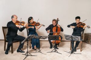 The Takacs Quartet, one woman and three men, sit in a row and perform their instruments.