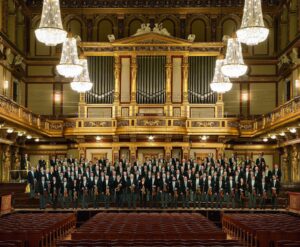 A wide group photo of the Vienna Philharmonic Orchestra, standing in front of an ornate golden organ, with dazzling chandeliers suspended above them.