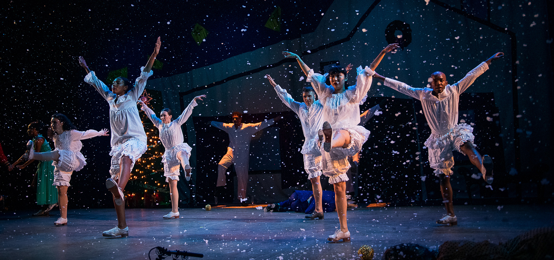 Dorrance Dance performers wear white costumes, and extend their arms upwards while snowflake-like specks rain down around them on the stage.