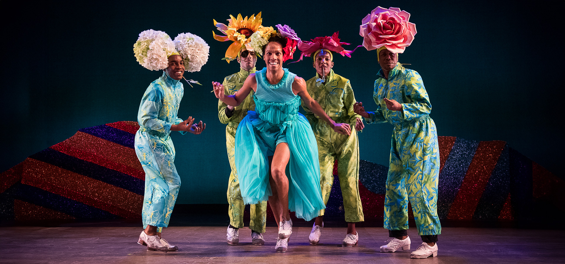 Performers in the Dorrance Dance group wear large flower headpieces and configure together in a tap-dance routine on stage.