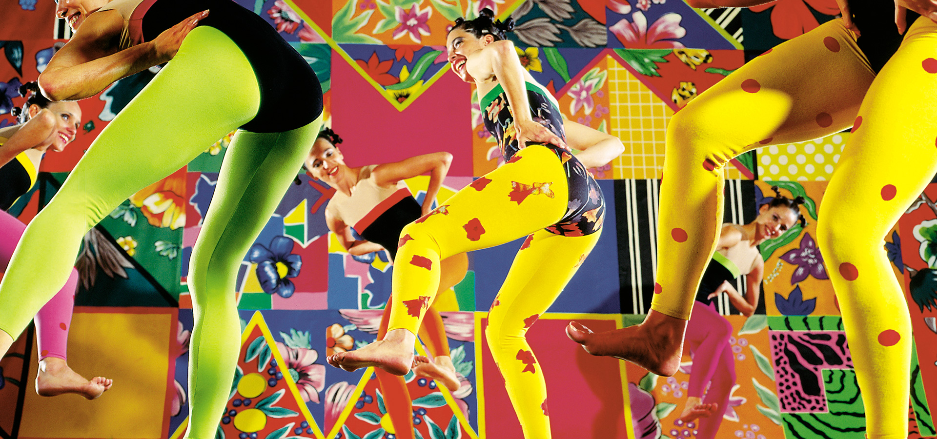 Dancers in the Grupo Corpo group wearing yellow leggings with black leotards, dancing against a vibrant, multi-patterned backdrop.