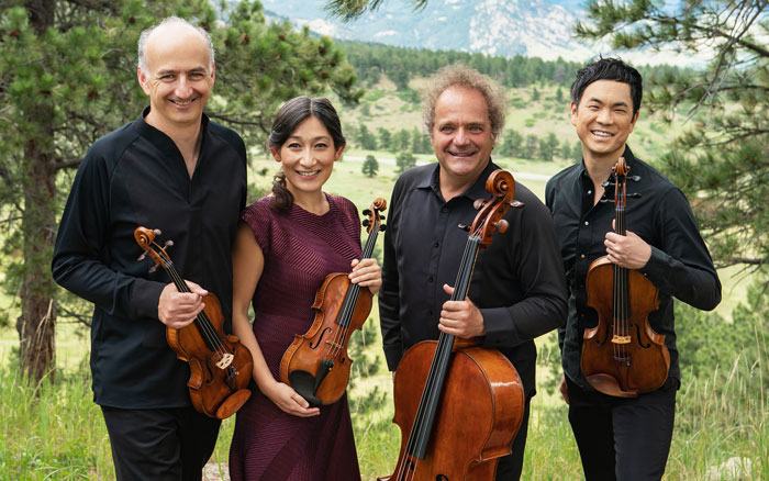 The Takács Quartet members, three men and one woman, stand smiling with their instruments in hand, photographed in the Colorado Mountains.