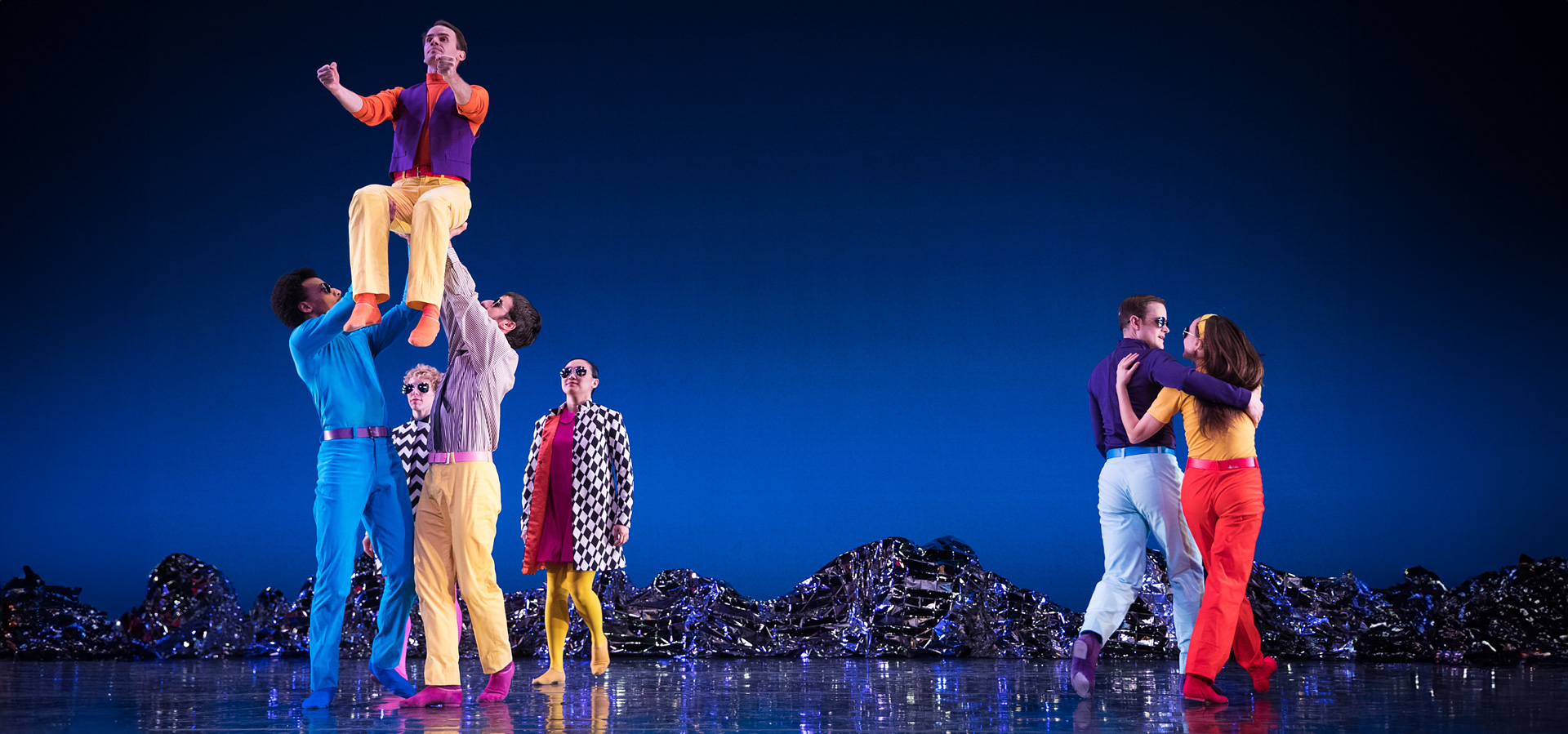 Dancers from the Mark Morris Dance Group perform Pepperland in colorful costumes on a blue-lit stage.
