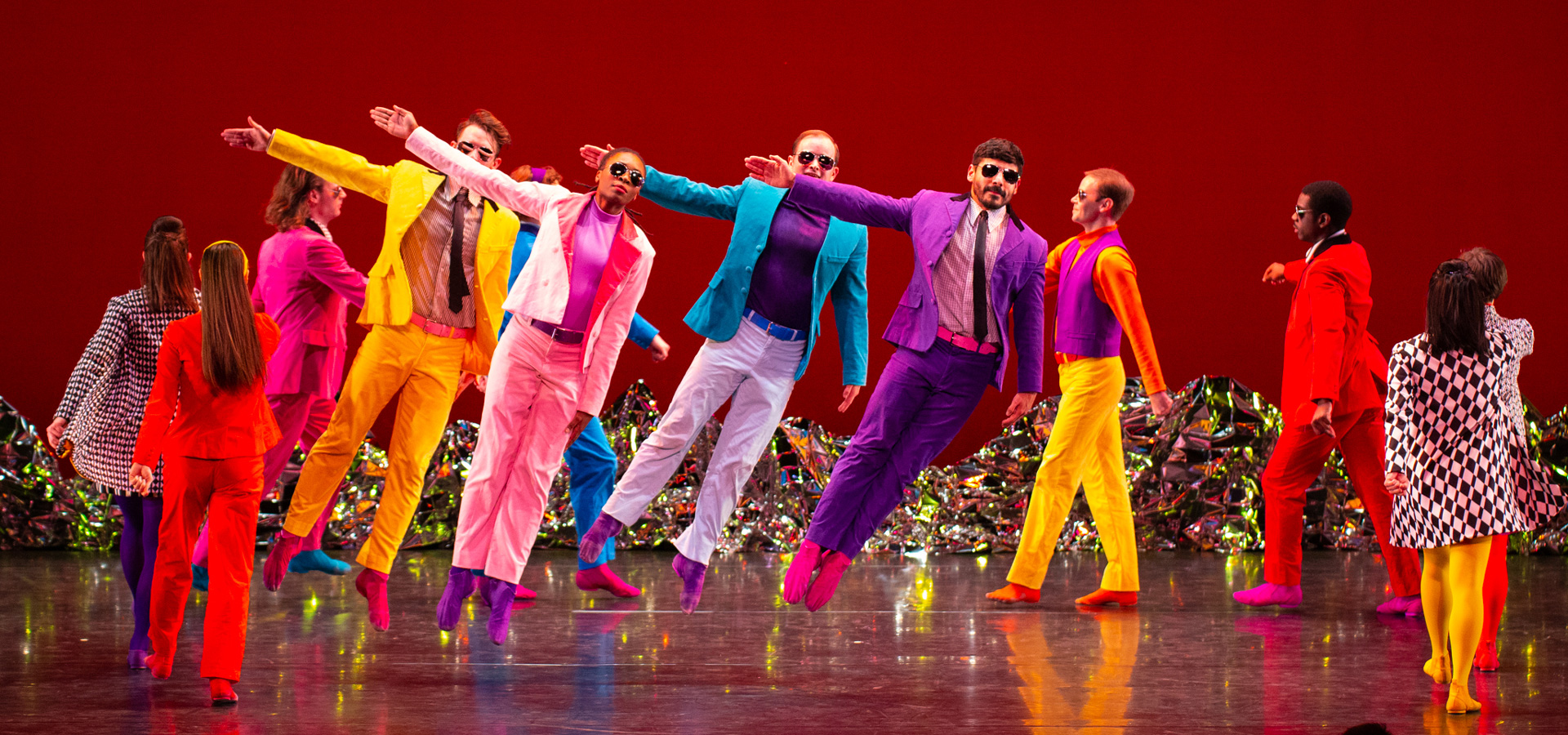Dancers performing Pepperland jump in the air with their arms extended, wearing neon colored suits.
