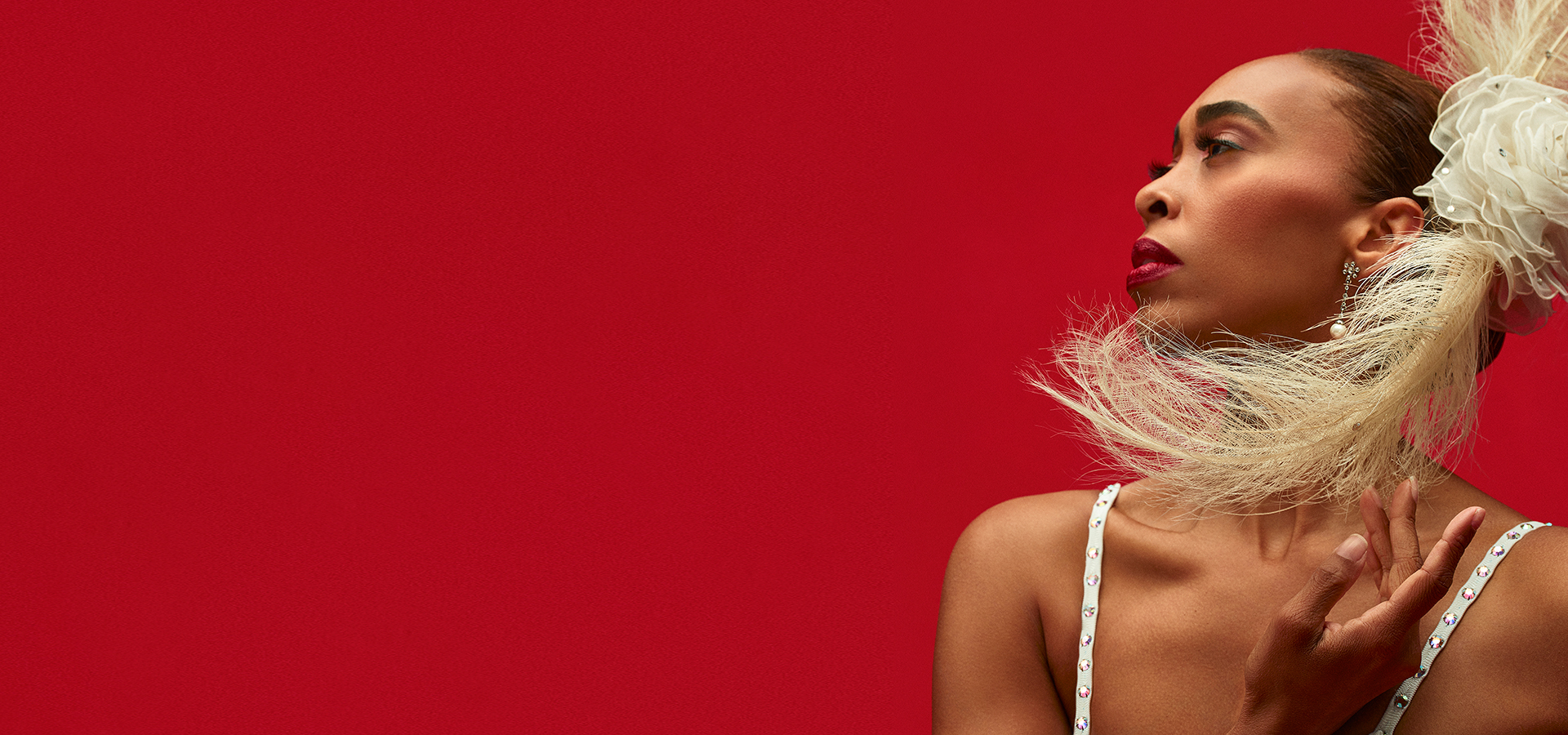 An Alvin Ailey American Dance Theater performer looking to the right, with a large feathered headpiece in her hair, standing against a bright red backdrop.