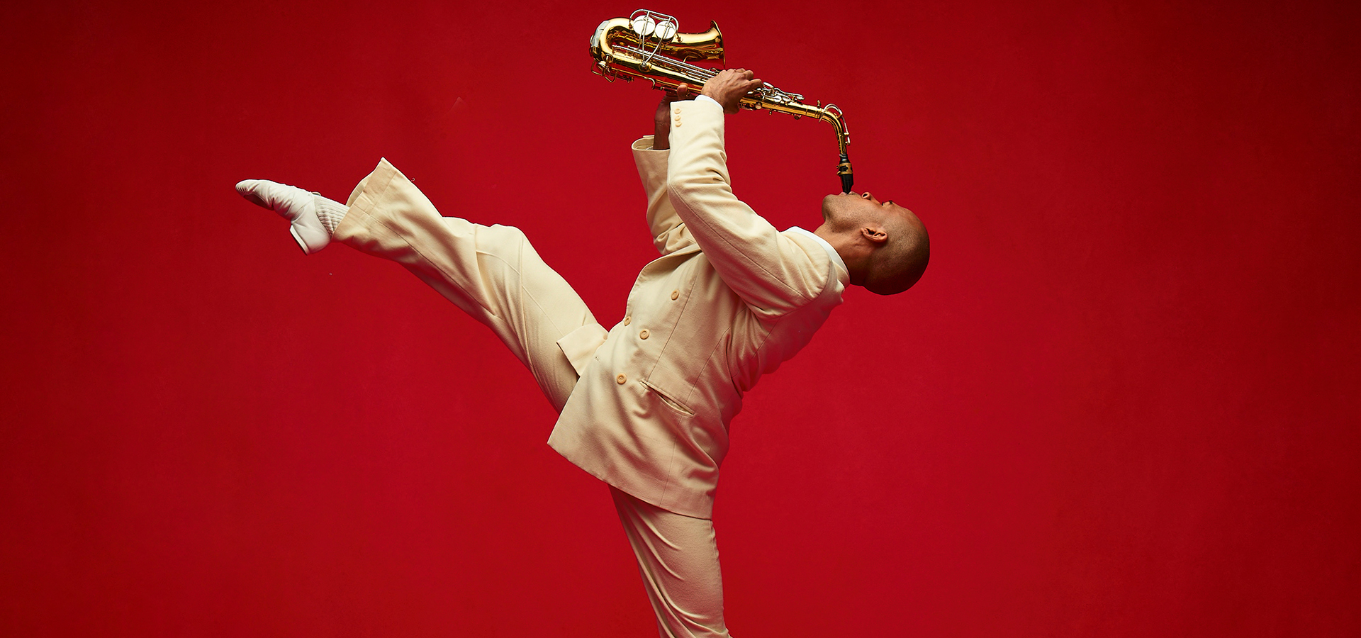 An Alvin Ailey American Dance performer in a cream costume, playing the saxophone with his leg lifted in an attitude.