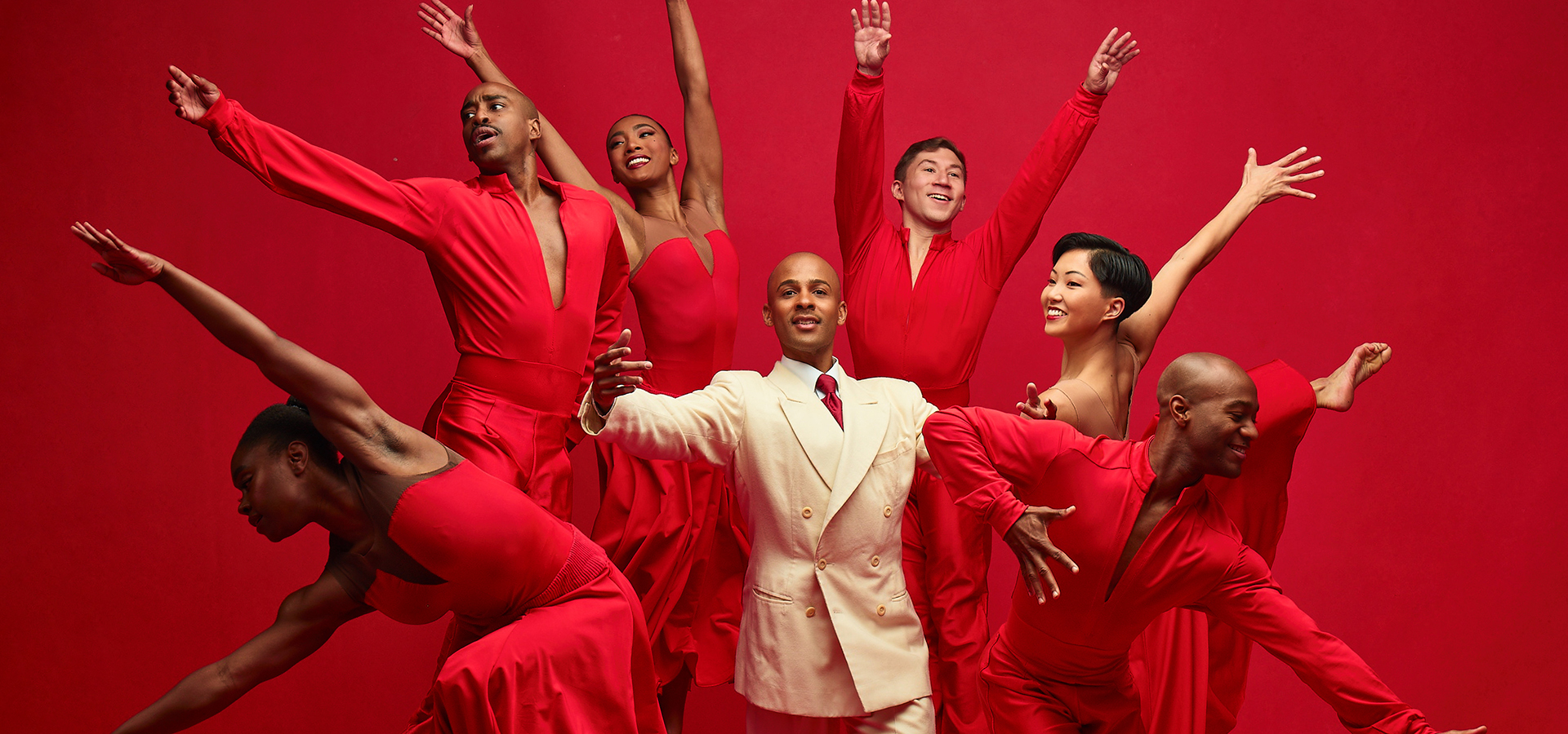 Ailey performers in red costumes with their arms raised surround a male dancer in a white suit posing in the center.