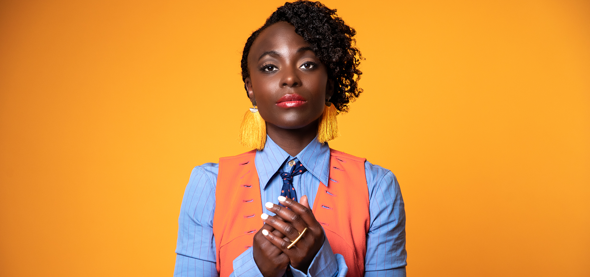 A vibrant portrait photo of artist Nathalie Joachim, a young black woman wearing a polka dot neck tie, and orange vest and orange tassle earrings.
