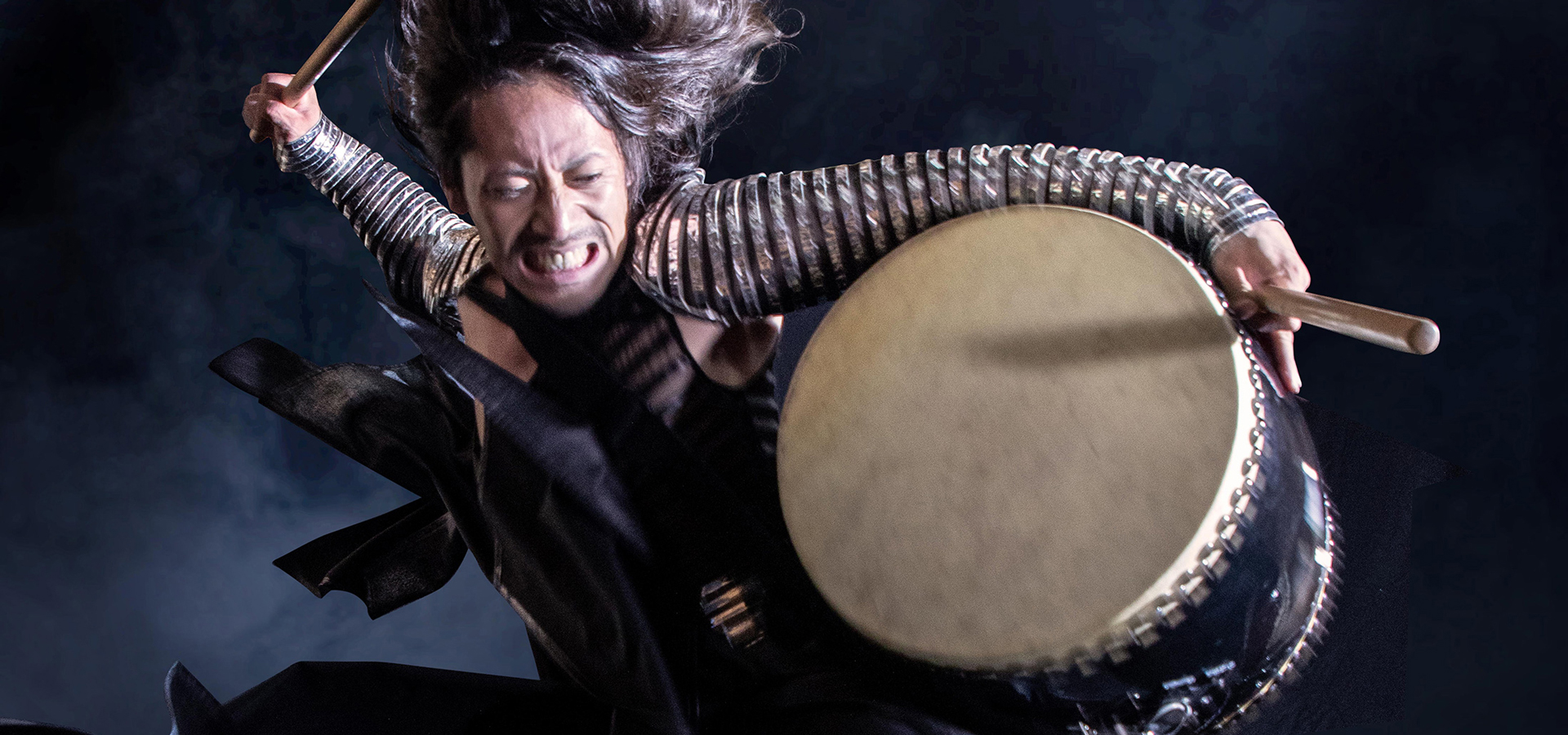A close up image of a long-haired Drum Tao performer rearing his arm to beat the large drum that he holds under his left shoulder, wearing elaborate silver arm bands.