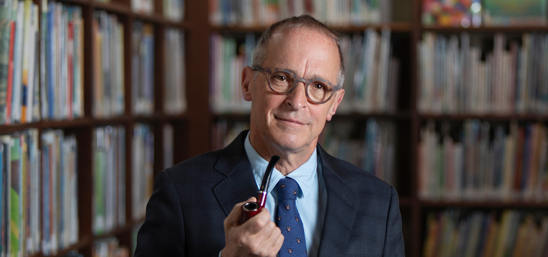 David Sedaris holding a pipe while wearing a blue suit and tie, standing in front of a wall of bookshelves.