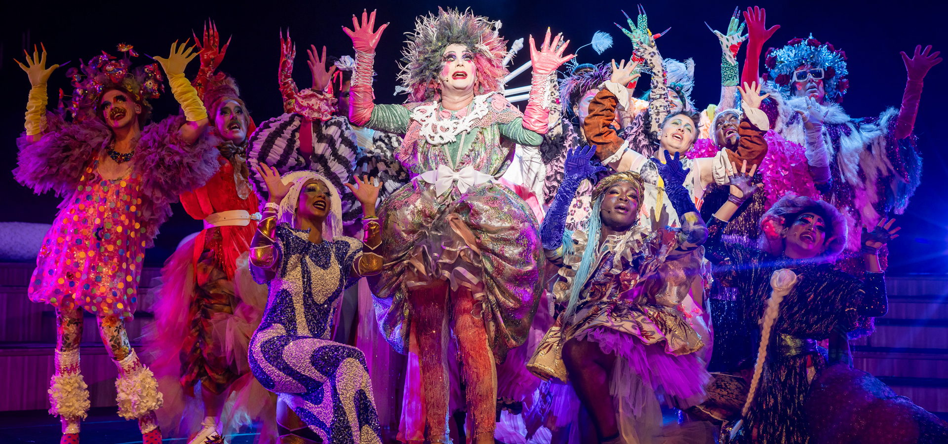 The bark of millions performers wear elaborately textured neon costumes and wigs, posing in a group on stage with their hands raised.