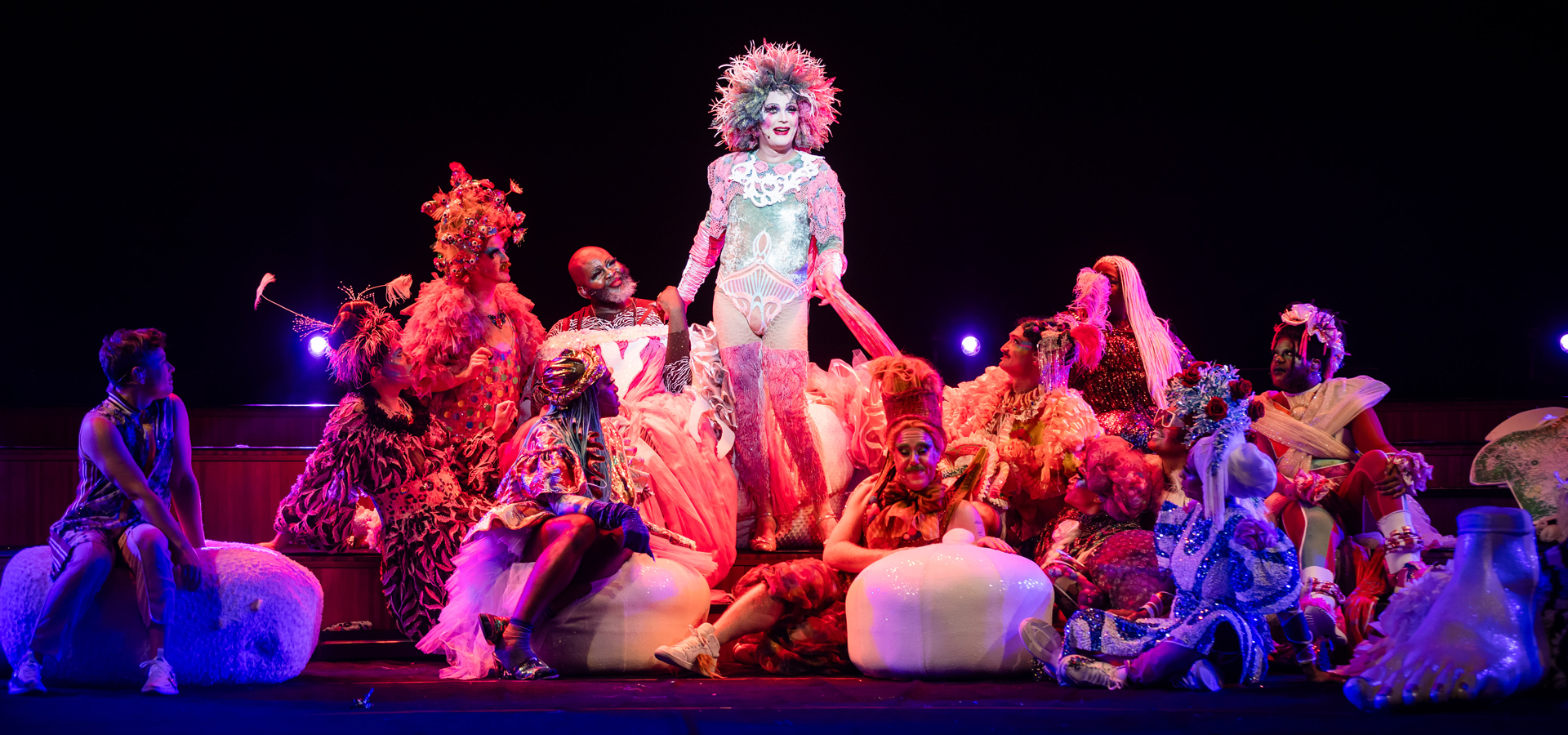 The bark of millions performers wear elaborately textured neon costumes and wigs, posing in a group on stage.