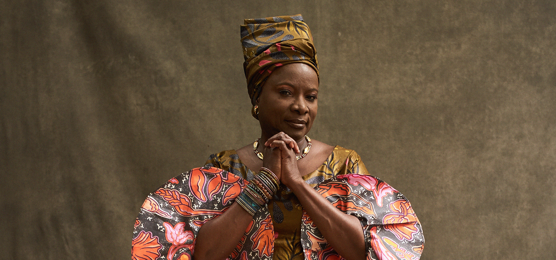 Angelique Kidjo wearing a patterned headwrap matching her dress. She sits with her hands clasped together, wrists adorned with a stack of bangle bracelets, and looks at the camera.