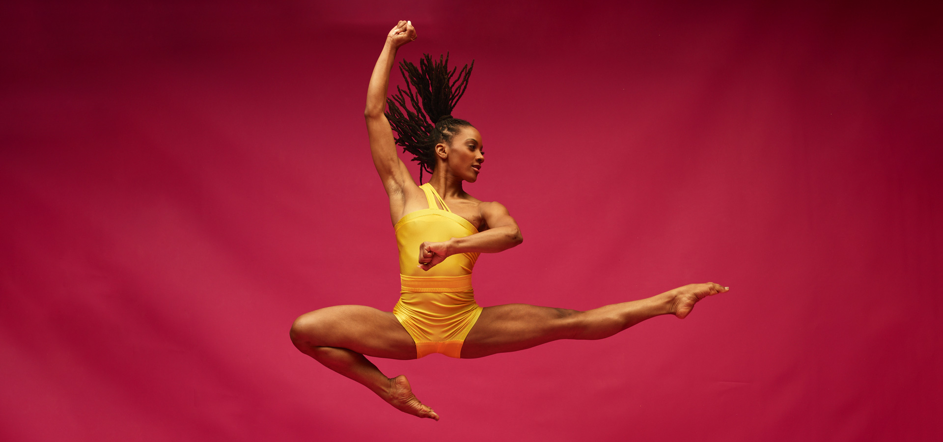 Dancer from the the Alvin Ailey American Dance Theater poses against a red background in a powerful jump with her arms and legs extended mid-air, wearing a vibrant yellow leotard.