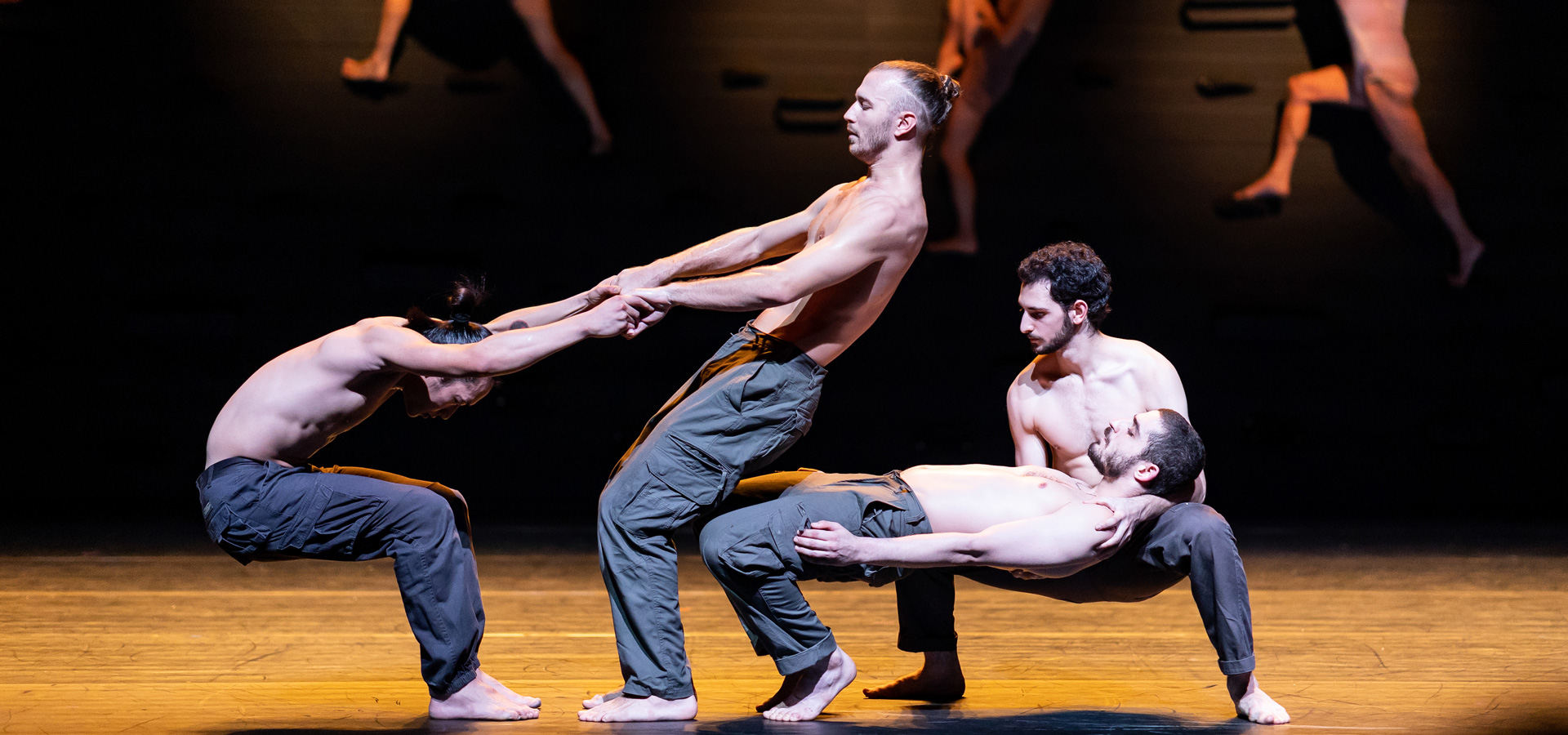 Four bare-chested male dancers in the Batsheva Dance group balance upon one another in an on-stage performance of MOMO.