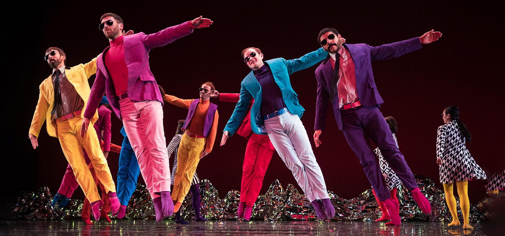 Dancers from the Mark Morris Dnace Group appear mid-jump in an on-stage performance, wearing vibrant attire with sunglasses over their eyes.