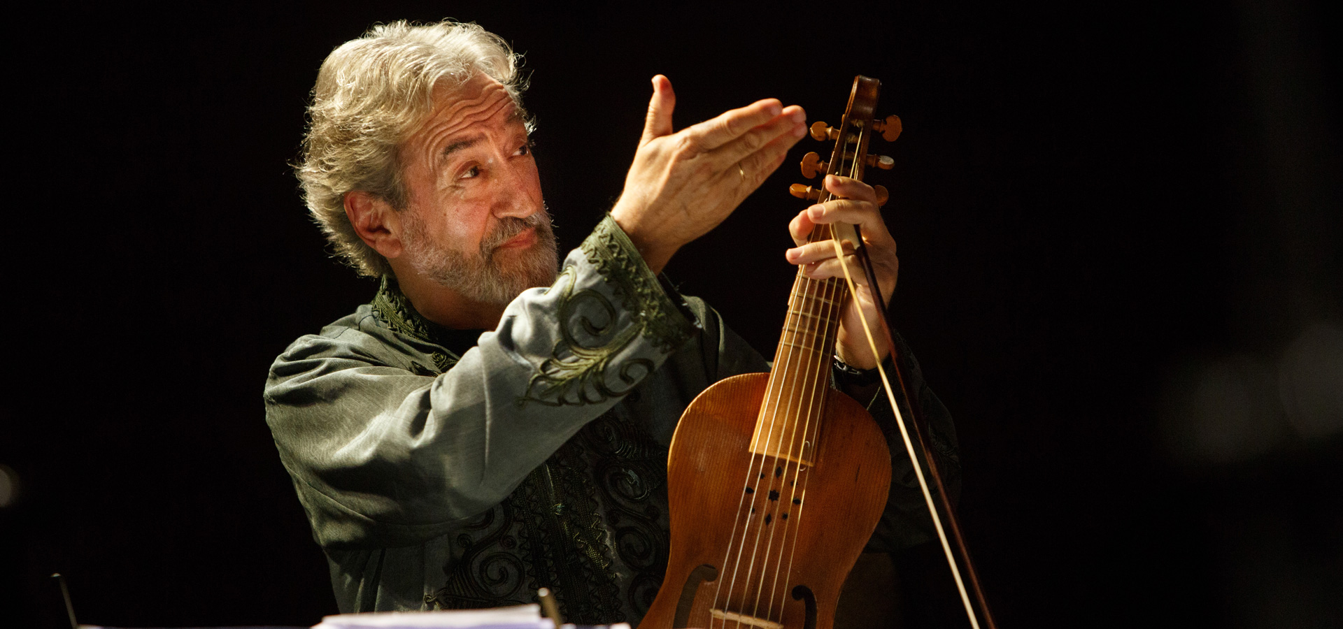 Spanish conducter and composer Jordi Savall shown against a dark background, holding his viol in one hand and raising his other in a gesture to the audience.