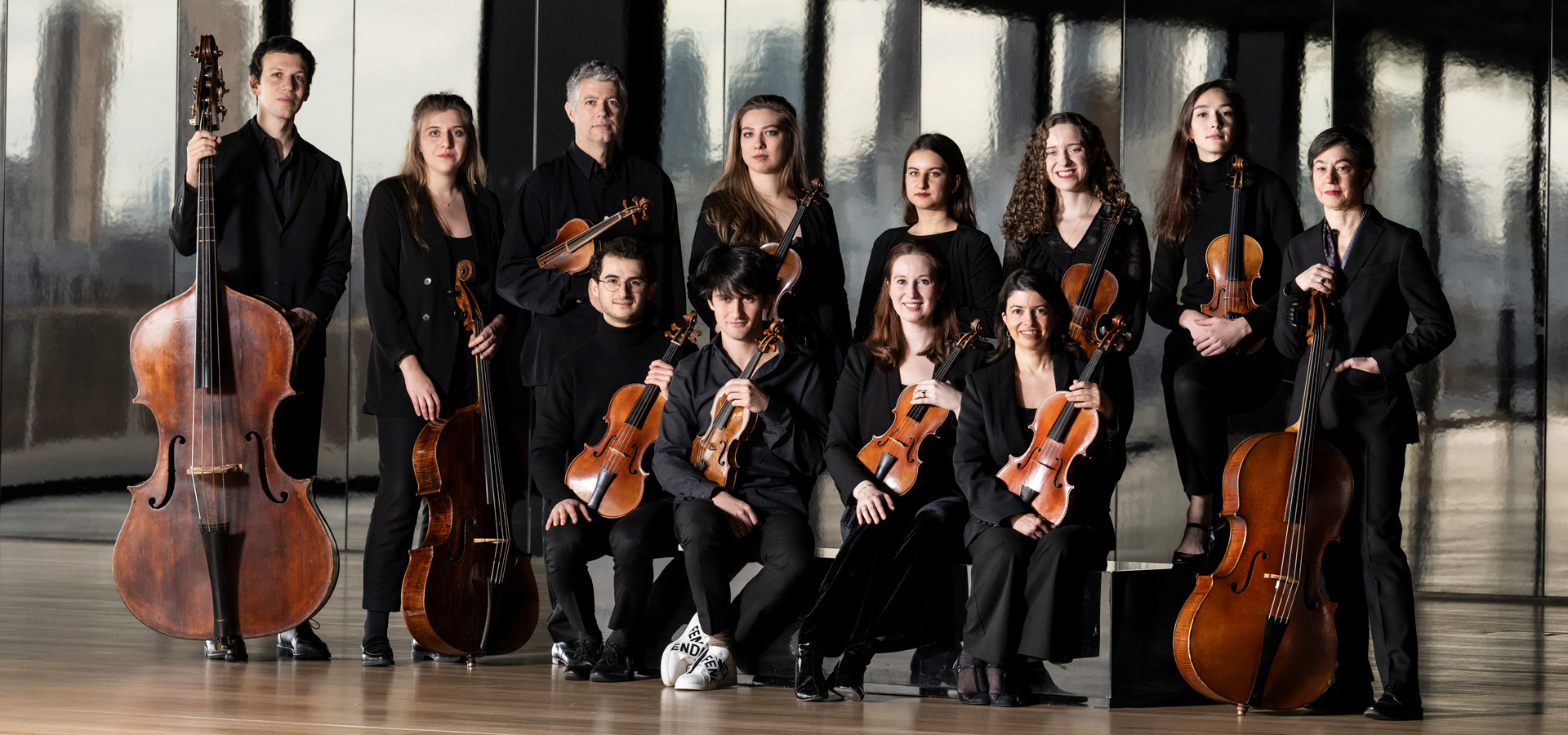 The twelve musicians of the Les Arts Florissants group wear black formal dress and pose with their instruments against a metallic backdrop.