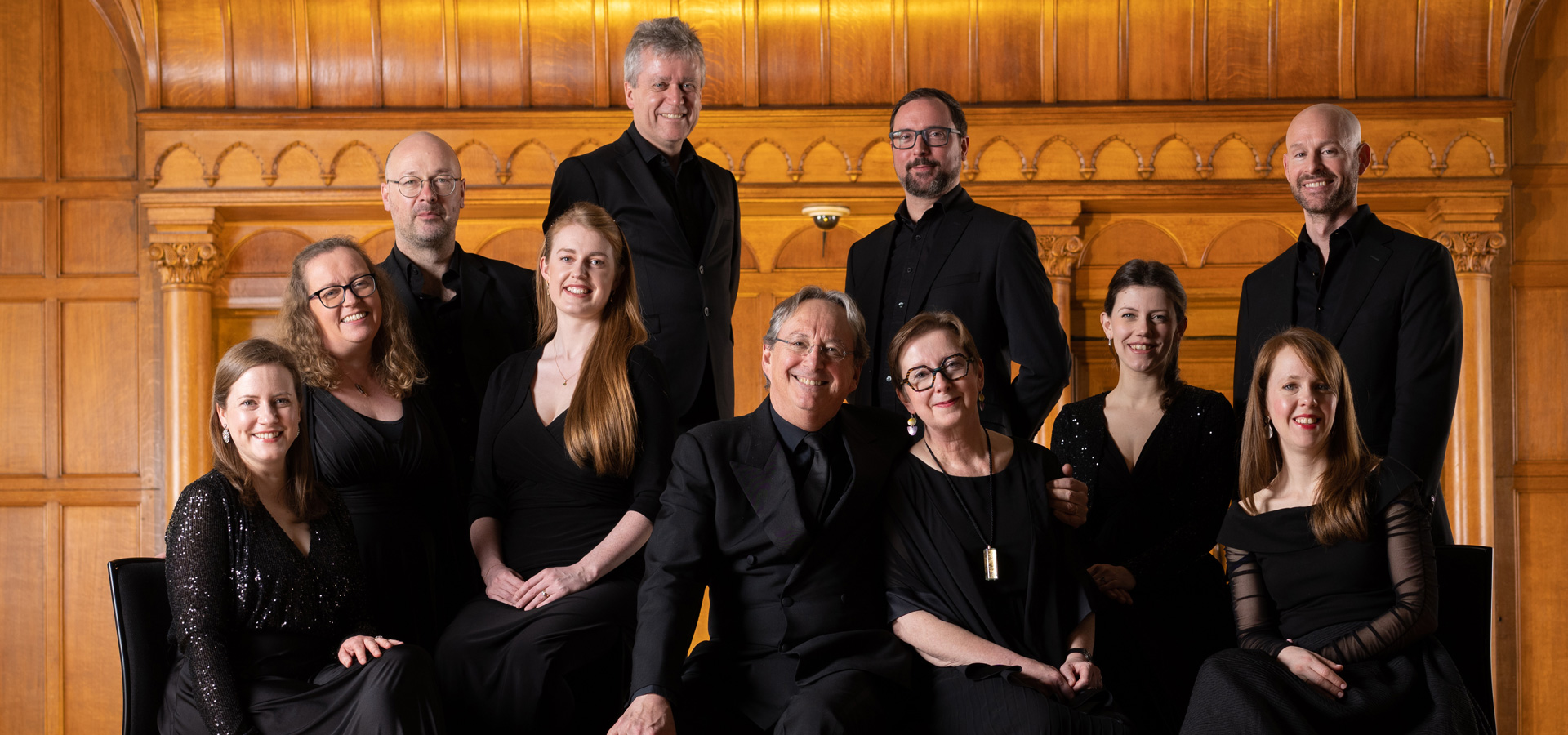 The Tallis Scholars smile happily at the camera while wearing black formal clothing, posing as a group in front of a gold wooden mantle.