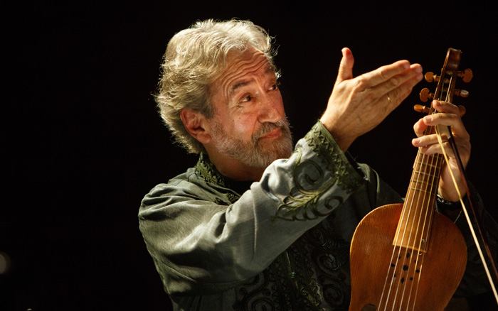 Spanish conducter and composer Jordi Savall shown against a dark background, holding his viol in one hand and raising his other in a gesture to the audience.