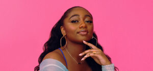 Samara Joy, a young black woman with long wavy hair wears a bright blue dress and overshirt that contrasts vividly with the hot pink background.