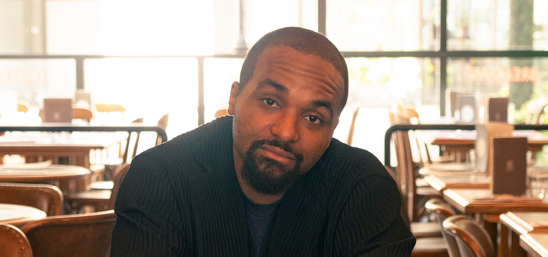 Sullivan Fortner looks directly into the camera while sitting backwards on a wooden chair and wearing a striped suit for this close up image.