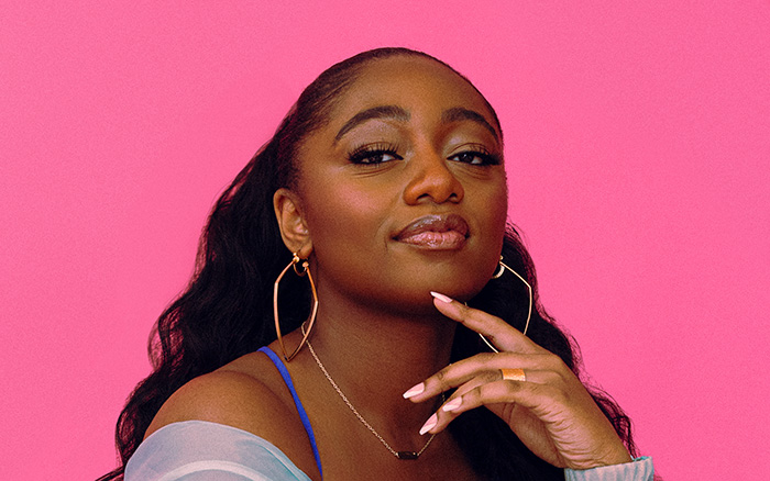 Samara Joy, a young black woman with long wavy hair wears a bright blue dress and overshirt that contrasts vividly with the hot pink background.