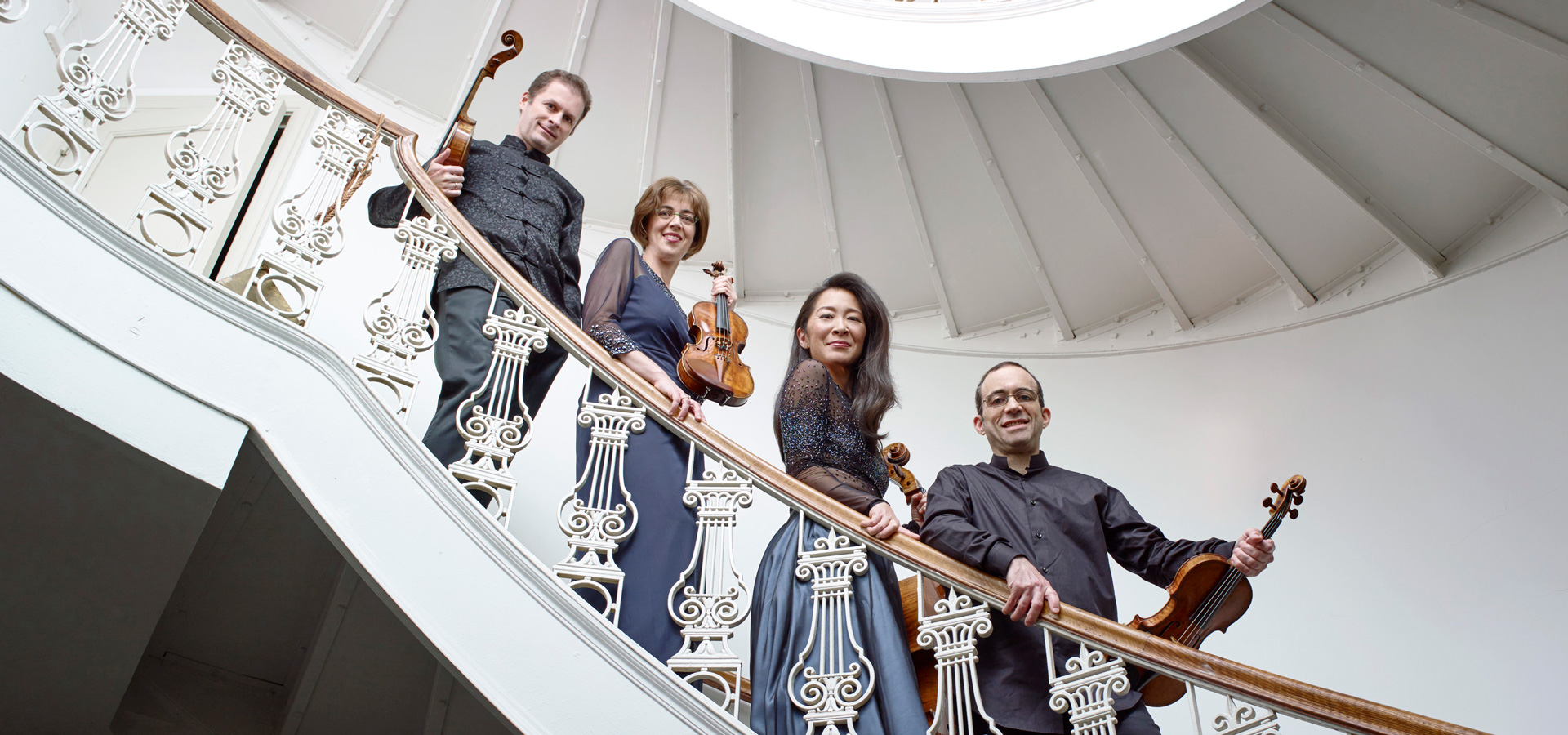 The Brentano String Quartet, two men and two women, hold their instruments and smile down at the camera from a white spiral staircase.