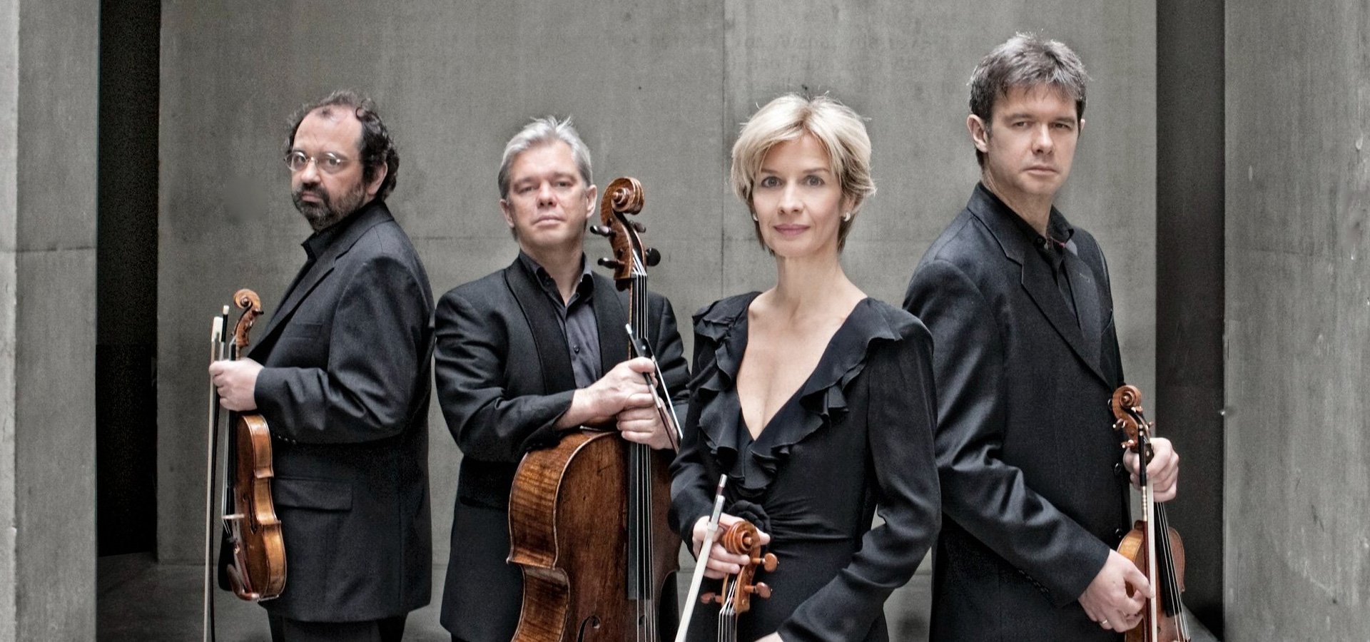 The three men and one woman of the Hagen Quartet stand in front of a concrete background, wearing black formal dress and holding their instruments at waist level.