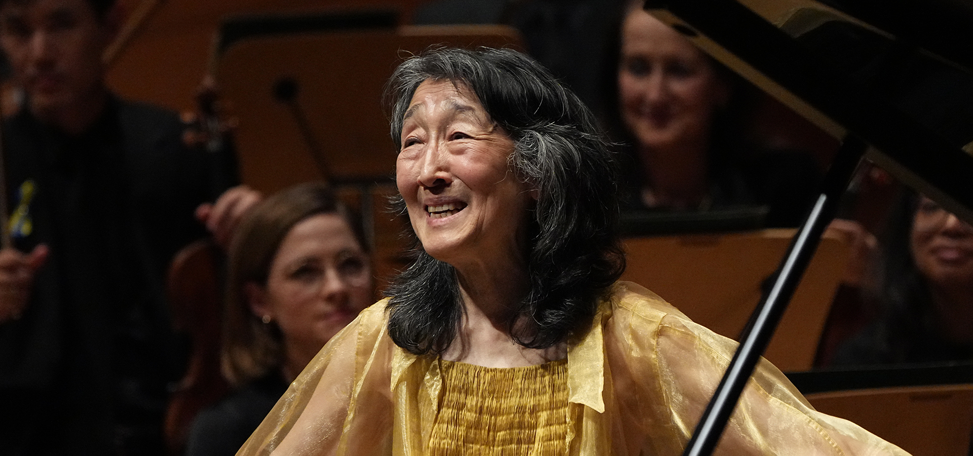 Pianist Mitsuko Uchida stands smiling in sparkling yellow attire, in front of her piano with the Mahler Chamber Orchestra behind her.