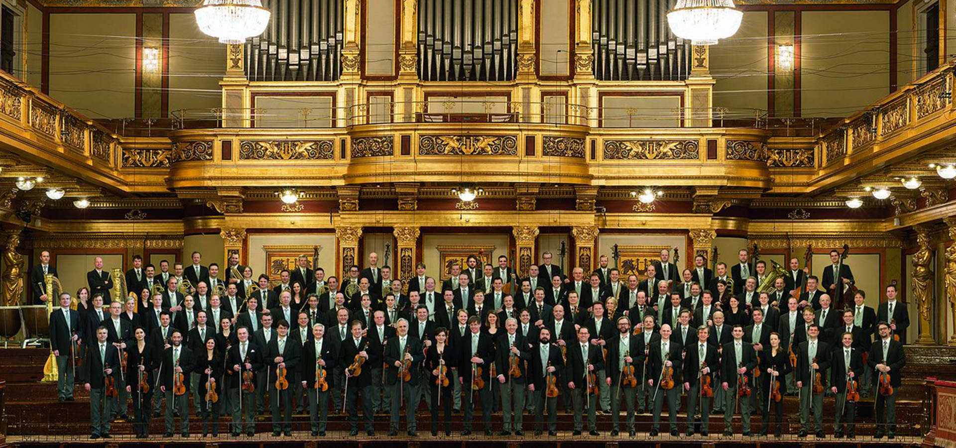 The Vienna Philharmonic stand as a large group in front of an ornate gold organ, holding their instruments and wearing formal dress.